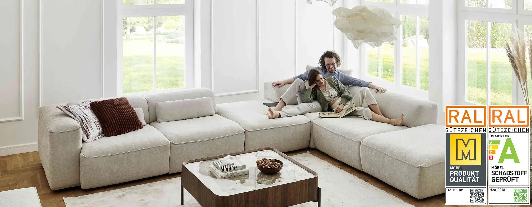 Großzügiges, helles Wohnzimmer mit bodentiefen Fenstern und modernem, beigem Ecksofa. Ein Paar sitzt entspannt auf dem Sofa und genießt gemeinsam ein Buch.